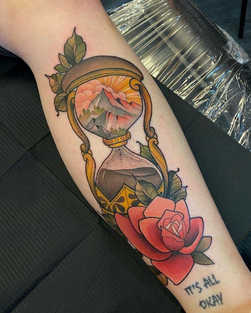 
"Neo-traditional tattoo featuring an hourglass with a mountain landscape inside, adorned with a vibrant red rose and leafy accents, complete with the phrase 'It's all okay', symbolizing the passage of time and natural beauty."